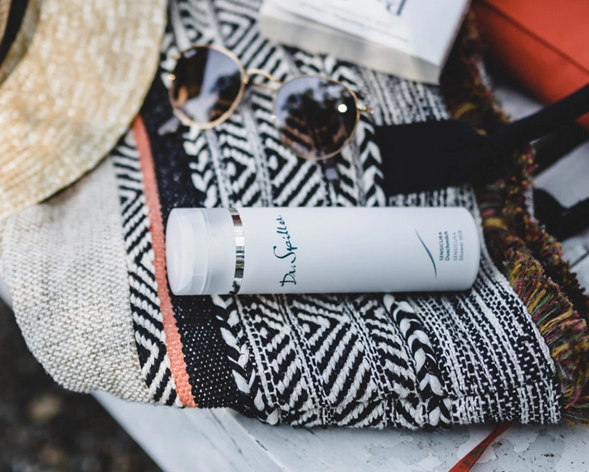 Sensicura Duschmilch Flasche liegt auf einem Badetusch neben einer Sonnenbrille